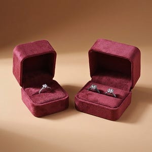 May include: Two open, deep red velvet ring boxes. The box on the left holds one silver-toned ring with a clear stone. The box on the right holds two silver-toned rings, each featuring a clear stone. The boxes have a square shape and soft texture.