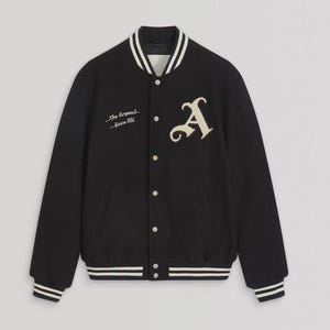 Può includere: Una giacca varsity nera con una lettera A color crema sul petto sinistro e il testo ricamato The Arsenal Since 1886 sul petto destro. La giacca presenta una chiusura frontale con bottoni a pressione e bordi a coste a righe bianche su colletto e polsini.
