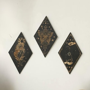 Puede incluir: Tres obras de arte murales en forma de diamante con diseños celestiales. Cada panel negro presenta ilustraciones doradas de una mujer, una mariposa y manos sosteniendo un sol y una luna. El arte está montado en una pared blanca.
