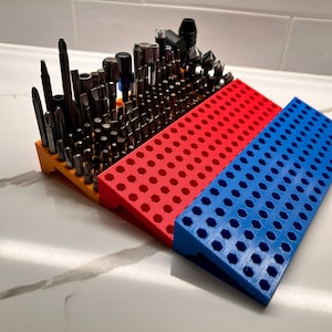 May include: A collection of black screwdriver bits and attachments in a multi-coloured holder. The holder has orange, red, and blue sections with numerous holes to hold the bits. The bits are of different sizes and shapes.