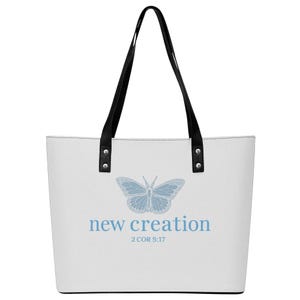 Puede incluir: Bolso tote blanco con asas negras. El bolso presenta un diseño de mariposa azul claro y las palabras "new creation" con la referencia bíblica 2 COR 5:17 en azul claro. Las asas están sujetas con remaches negros.