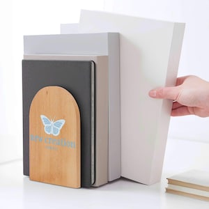 Puede incluir: Un sujetalibros de madera con una mariposa azul claro y la inscripción "new creation". Sostiene varios libros de diferentes tamaños y colores. Una mano sostiene un libro blanco.