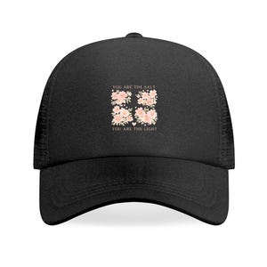 May include: Black trucker hat with a curved brim and mesh back. The front features the text "YOU ARE THE SALT YOU ARE THE LIGHT" above a floral design with pink flowers and green leaves. A stylish accessory.