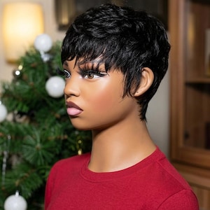 May include: A mannequin head featuring a short, layered black pixie cut wig with textured bangs. The mannequin has brown eyes, long dark eyelashes, white eyeliner, and light pink lips, wearing a plain red crew neck shirt.