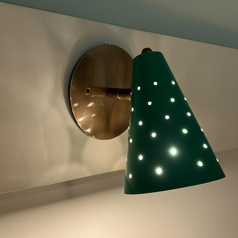 Puede incluir: Aplique de pared verde en forma de cono con perforaciones de puntos blancos, montado sobre una base circular de color lat&oacute;n. El aplique tiene un brazo de lat&oacute;n y est&aacute; fijado a una pared blanca.