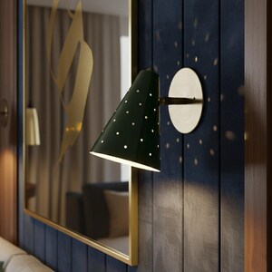 Puede incluir: Aplique de pared verde oscuro con una pantalla c&oacute;nica salpicada de peque&ntilde;as perforaciones blancas. El aplique est&aacute; montado sobre una base y un brazo circulares de color lat&oacute;n, que iluminan una pared azul oscuro. Un gran espejo con marco dorado en el fondo.