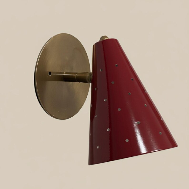 Puede incluir: Aplique de pared c&oacute;nico rojo con peque&ntilde;as perforaciones circulares. Cuenta con una placa de montaje y un brazo de color lat&oacute;n. El aplique est&aacute; montado en una pared clara.