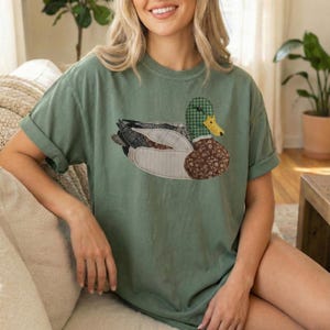 Könnte beinhalten: Salbeigrünes T-Shirt mit einem Enten-Applikation. Das Enten-Design verwendet verschiedene Stoffmuster, darunter Karos, Blumenmuster und unifarbene Farben. Das T-Shirt hat hochgekrempelte Ärmel und eine lockere Passform.