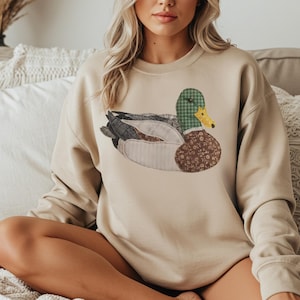 May include: A beige sweatshirt with a patchwork mallard duck design. The duck has a green and yellow checked head, a brown floral patterned body, and gray and white wings. The sweatshirt has long sleeves and a crew neck.