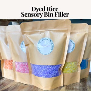 May include: Several brown paper bags filled with dyed rice in various colors, including orange, pink, purple, yellow, and blue. Each bag has a clear window showing the rice and a circular sticker that reads "PLAY TO LEARN CREATIONS". The text "Dyed Rice Sensory Bin Filler" is at the top.