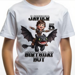 Puede incluir: Camiseta blanca con una ilustración de dibujos animados de un niño montando un dragón negro. El texto "JAVIER 5 BIRTHDAY BOY" está impreso en plata, con contorno. El niño lleva un atuendo marrón y verde.