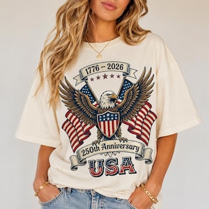 Puede incluir: Una camiseta de manga corta color crema con un gráfico de un águila calva con las alas extendidas, dos banderas estadounidenses y el texto 1776-2026, 250th Anniversary y USA. El diseño incluye los colores rojo, blanco y azul con cinco estrellas.