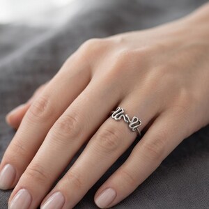 Könnte beinhalten: Ein silberner Ring mit einem durchgehenden, fließenden Drahtdesign, das zwei miteinander verbundene, abgerundete Schlaufen an der Oberseite bildet. Das Metall hat eine glatte, matte Oberfläche und der Ring ist dünn und gleichmäßig geformt.