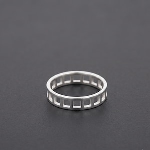 May include: A silver-coloured ring featuring a continuous band design composed of a series of evenly spaced, rectangular cutouts that create a grid-like pattern around the entire circumference of the circular metal band.