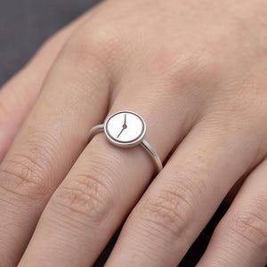 May include: A silver-toned ring featuring a circular face designed to resemble a clock dial. The centre of the round setting displays two thin, dark hands positioned against a plain, light-coloured background, mimicking the appearance of a watch face.