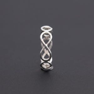 Könnte beinhalten: Ein silberner Ring mit einem durchgehenden Band aus miteinander verbundenen Unendlichkeitssymbolen. Das Metall hat eine polierte, reflektierende Oberfläche und das Design besteht aus einem sich wiederholenden Muster aus Schleifen.