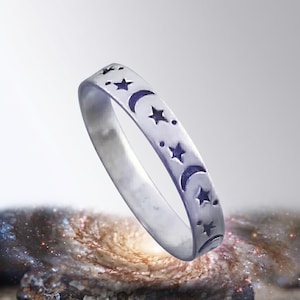 May include: A silver-toned metal ring featuring a repeating pattern of engraved crescent moons and five-pointed stars. The band has a smooth, polished finish with dark, recessed details that highlight the celestial motifs across the outer surface.