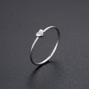 Könnte beinhalten: Ein dünner, polierter Ring aus silberfarbenem Metall mit einem kleinen, glatten herzförmigen Anhänger in der Mitte des Bandes. Der Ring hat ein schlichtes, kreisförmiges Design mit einer reflektierenden Oberfläche und einem gleichmäßigen Finish.