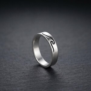 Peut inclure: Une bague en métal argenté avec une finition lisse et polie, ornée d'un motif de vague noire gravé qui s'enroule partiellement autour de l'anneau extérieur. La bague présente un profil arrondi et une largeur constante sur toute sa structure.