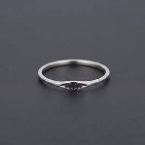 Puede incluir: Un anillo fino de metal plateado pulido que presenta un pequeño emblema grabado con forma de murciélago centrado en la parte frontal de la banda. El anillo tiene un perfil liso y redondeado con un acabado metálico que refleja la luz.
