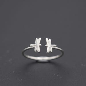 May include: A silver-toned open ring featuring two small dragonfly motifs facing each other at the center. The band is thin and circular, with the dragonfly wings extending outward to create a gap between the two insect designs on the top.