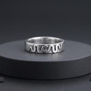 May include: A silver-coloured metal ring featuring a continuous, embossed snake design that wraps around the entire outer band. The surface of the ring has a polished finish with darkened recessed areas that highlight the detailed serpent motif.