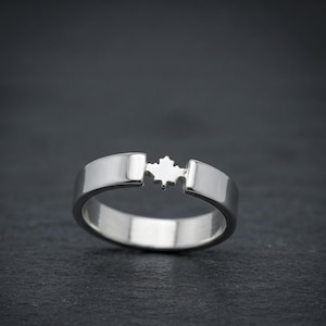 May include: A polished silver-toned ring featuring a smooth, solid band that is interrupted at the top by a small, centered cutout in the shape of a maple leaf. The metal has a reflective, metallic finish and a clean, continuous circular design.