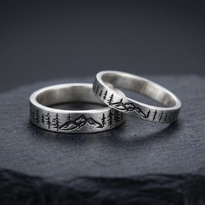 May include: Two silver rings featuring a continuous engraved design of a mountain range and pine trees. The metal has a polished finish with darkened, recessed details that highlight the landscape pattern encircling the band of each ring.