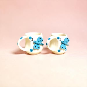 Può includere: Due piccole tazze in ceramica color bianco sporco con una decorazione a libellula blu in rilievo sulla parte anteriore. Le tazze sono decorate con piccoli pois blu sparsi sulla superficie, compresi i manici curvi.