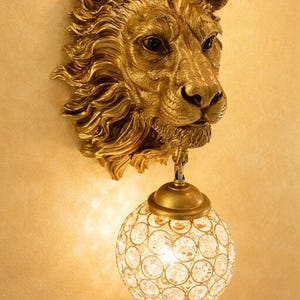 May include: A wall-mounted light fixture featuring a sculpted gold-colored lion head with a detailed mane. Suspended from the lion's chin is a spherical lamp shade constructed from a gold metal lattice frame embedded with clear glass crystals.