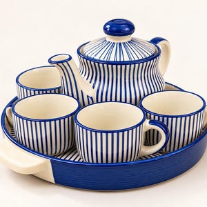 Può includere: Un set da tè in ceramica composto da una teiera con coperchio e quattro tazze abbinate, tutte decorate con strisce blu verticali su fondo crema. Il set è disposto su un vassoio da portata rotondo bicolore con un bordo blu solido.