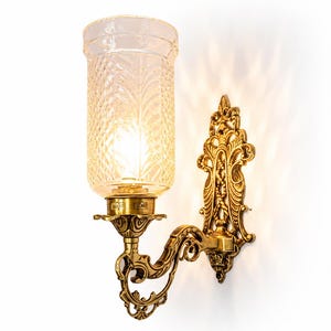 Pu&ograve; includere: Una lampada da parete con una piastra di montaggio in ottone lucido ornata e un braccio decorativo curvo. L'apparecchio sostiene un paralume cilindrico in vetro trasparente con una superficie testurizzata a rombi che diffonde la luce.