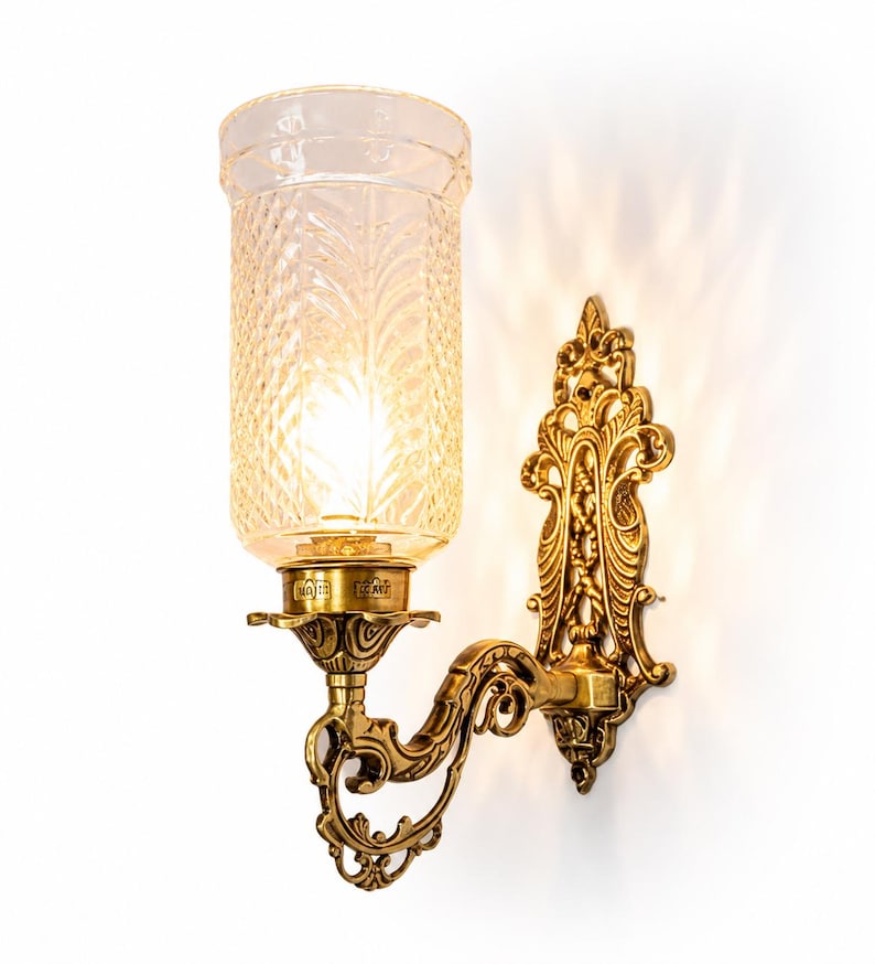 Pu&ograve; includere: Una lampada da parete con una piastra di montaggio in ottone lucido ornata e un braccio decorativo curvo. L'apparecchio sostiene un paralume cilindrico in vetro trasparente con una superficie testurizzata a rombi che diffonde la luce.