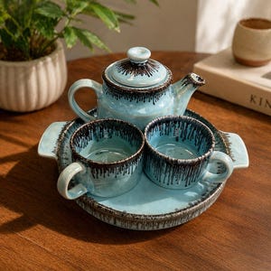 Puede incluir: Juego de té de cerámica azul claro con detalles marrón oscuro. El juego incluye una tetera, dos tazas y una bandeja. El juego de té está sobre una mesa de madera. Una pequeña planta y un libro son visibles en el fondo.