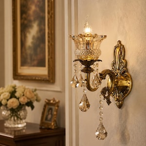 May include: A wall-mounted light fitting featuring an antique brass finish backplate with ornate scrollwork. A clear, fluted glass shade holds a single candle-style bulb, accented by multiple hanging clear crystal teardrop prisms and beads.