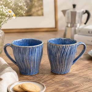 May include: Two ceramic mugs featuring a vibrant blue glaze with a vertical ribbed texture and a slightly flared rim. Each mug has a sturdy handle and a rustic finish, showcasing a deep blue color with lighter highlights along the raised ridges.