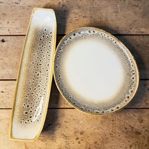 May include: A set of three ceramic mugs with a speckled, matte finish. The mugs are in shades of cream, light gray, and dark gray. Each mug has a simple, minimalist design and a rounded handle.