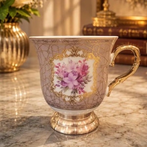 Può includere: Una tazza in porcellana con uno sfondo malva tenue e un motivo geometrico dorato. Il centro presenta un bouquet floreale nei toni del rosa e del viola, incorniciato da un bordo dorato decorato. È dotata di un manico e una base dorati.