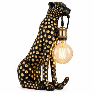 Puede incluir: Una lámpara de mesa con forma de leopardo sentado con acabado negro mate y manchas circulares doradas. El leopardo sostiene en su boca un casquillo de color latón, que cuenta con una bombilla grande, redonda y de cristal transparente.
