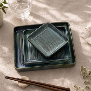Può includere: Un set di tre piatti quadrati in ceramica di diverse dimensioni, caratterizzati da uno smalto reattivo blu e verde acqua profondo con una texture a coste verticali sulla superficie. I piatti sono impilati e accompagnati da bacchette in legno.