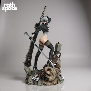 Puede incluir: Figura detallada de un personaje femenino del juego Nier: Automata. Está en una pose dinámica, sosteniendo una espada grande y una hoja más pequeña. La figura viste un atuendo negro con detalles blancos y se encuentra sobre una base rocosa. El texto "rath space" está en la esquina superior izquierda.