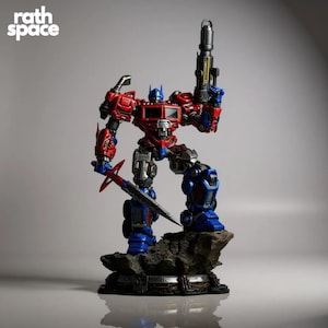 May include: An Optimus Prime figurine, predominantly red and blue, with silver and black detailing. The figure is posed on a rocky base, holding a gun and a sword. The "rath space" logo is visible in the top left corner.