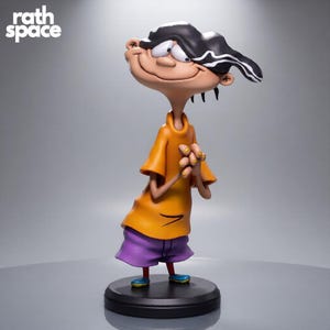 Puede incluir: Una figura coleccionable del personaje de dibujos animados Edd de Ed, Edd y Eddy, con cabello negro, una camiseta naranja y pantalones cortos morados. El personaje se encuentra sobre una base negra circular con el texto rath space visible arriba a la izquierda.