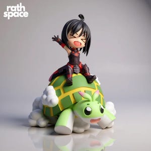 May include: A figurine of a cartoon character riding a green and yellow turtle. The character has black hair and is wearing a black and red outfit. The turtle has a smiling face and white cloud-like accents. The text "rath space" is in the upper left corner.
