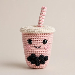 May include: A crocheted bubble tea plushie featuring a light pink cup with a cream-colored lid and a pink and white striped straw. The cup has black safety eyes, a stitched black smile, pink blush circles, and black crocheted boba pearls.