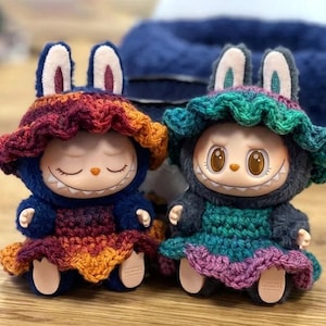May include: Two whimsical dolls, each adorned with a knitted bunny hat and matching outfit. One doll wears a blue and rainbow-colored ensemble, while the other is in teal and purple. Both have closed eyes and a wide, toothy grin.