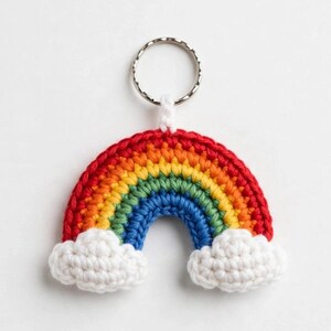 May include: A crochet keychain featuring a rainbow design with concentric bands of red, orange, yellow, green, and blue yarn. The rainbow is anchored by two white crocheted clouds at the base and is attached to a circular silver metal split ring.