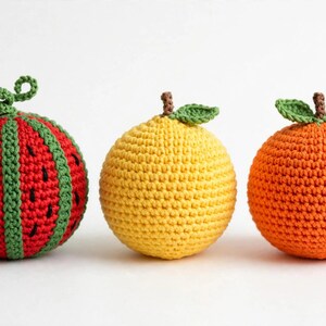 May include: Three spherical crochet toys shaped like fruit. The first is a red and green watermelon with black seed details and a curly stem. The second is a solid yellow lemon with a small green leaf. The third is a textured orange fruit.