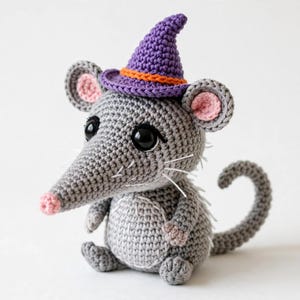 May include: A grey crocheted mouse plush toy featuring a pointed purple witch hat with an orange band, large black safety eyes, a small pink nose, pink inner ears, and a long curled tail. The mouse has white stitched whiskers and a round belly.