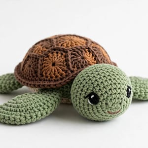 May include: A crocheted turtle toy featuring a light green head and flippers with a textured surface. The shell is constructed from dark brown and tan yarn in a hexagonal pattern. The turtle has black and white embroidered eyes and a small mouth.